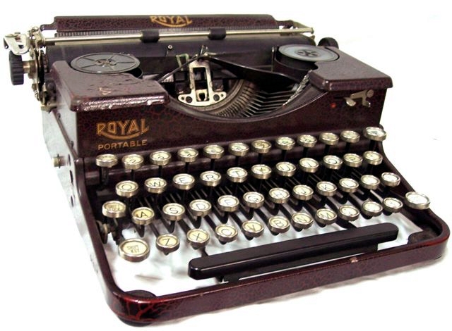 This photo of the left front quarter of the machine shows its flat-top design with recessed but visible ribbon spools, the paper bail with two rubber rollers, the Royal logo on the paper table and the right front of the case (with Portable beneath it), the left-hand carriage return, and the red crinkle paint. There are two platen twirlers. The keys are bright metal rimmed, with white background and black letters. The ribbon color selector is on the left front of the case.