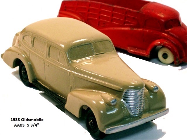 A gleaming, tan 1938 Oldsmobile 4-door sedan seen from the right front quarter. Like the '37, the car has suicide doors, white wheels, but is 5 3/4 inches long. Above it is a 1936 stake-side pickup truck made by Sun Rubber.