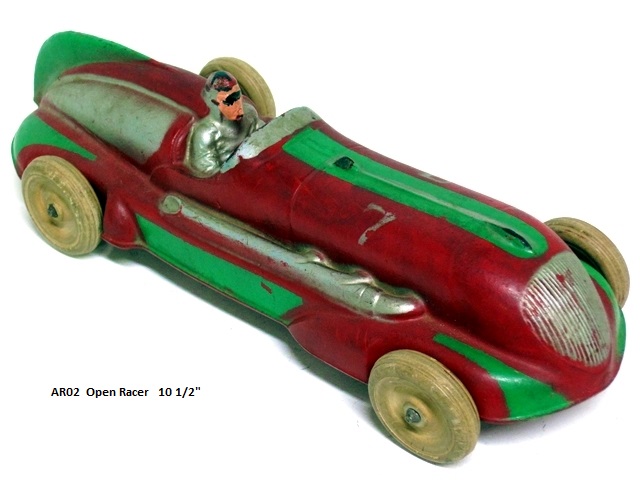 Front quarter view of the 10 1/2 inch long open racer.  With the somewhat spaceship design the race car is primarily red with silver on the front grill and on the both sides just behind the driver. The body of this racer is very aerodynamic with a green tail fin and two rounded equally aerodynamic fenders at front and rear. These have green, tear-shaped highlights, and the hood has a green highlight along the center line. The cockpit and driver are silver, and the wheels are white. The exhaust pipes come out from the hood (three to a side) and run along to the back edge of the cockpit on both sides of the race car. The number 7 is painted on the sides of the hood. The windscreen is silver, steering ring black, the dash red. A big, splendid toy!