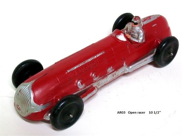 Front quarter view of the 10 1/2 inch open Indianapolis racer. The body is long and skinny, painted red with silver along the lower sides front and rear where the stabilizer bars are shown. There is also silver at the back of the cockpit. The silver exhaust runs along the right hand side. Auburn is lettered along the sides of the hood just above the silver colored hood louvers. The wheels, about an inch and a half in diameter, are black. The driver is wearing a silver top and helmet. He has flesh tone paint for his face and hands, and is wearing black goggles. The windscreen is silver.