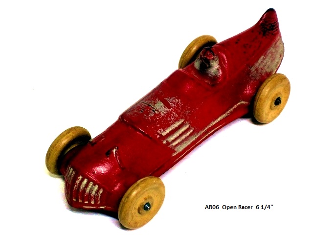 Front quarter view of the midsize 6 1/4 inch long open racer. This red race car has a low, wide, silver barred grill, the number seven on the side, silver horizontal louvers at the midpoint, the bottom one of which extends to the rear where it joins rounded, silver rounded ridges that sweep around the rounded back end of the body. The driver sits in a deep cockpit. There is a rounded fin at the midline of the body at the rear. The driver is silver with a red and black helmet. The white tires are large, about an inch and an eigth