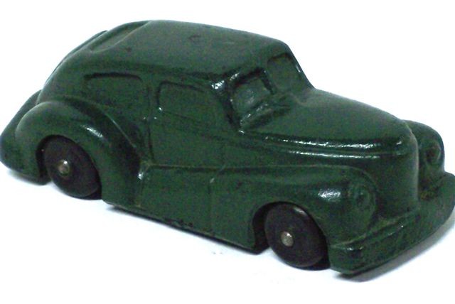 This is a front quarter view along the passenger side. The car is a deep green with black wheels. It is a fastback style coupe with a split windshield and a wide oval rear window. The fenders sweep downward from the front headlights to the middle of the door, then upward to the tail lights. The headlights are in the fenders, there is a wide radiator with horizontal bars, and a short, wide, stubby hood. The bumpers are wide and thick, and the car gives an overall impression of being a very sturdy toy.