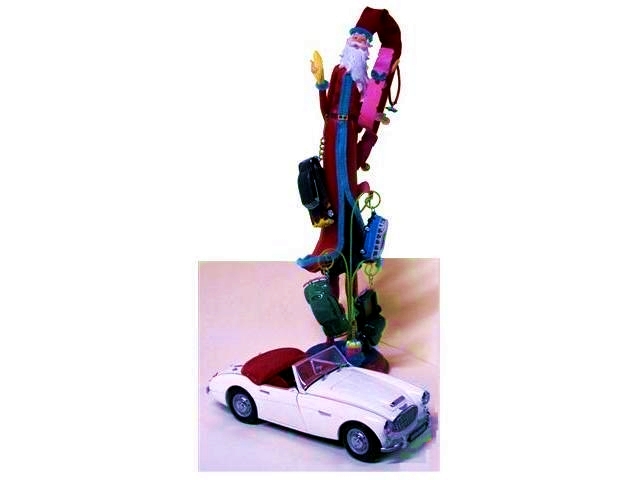 Pere Time s a Christmas tree ornament on a stand. A tall, skinny Father Time or Santa with a long white beard, and a long red overcoat with a bright blue trim. The green stand is akin to a paper spike and has four thin metal rings attached to leaf-like spurs along its length. From these rings depend Hotwheels toys, a hotrod, a VW Bus and VW Beetle, and a pickup truck. Sitting at the base of it is a 1:18 white Austin Healey roadster with red interior and steel wheels.