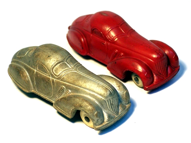 Pair of 1936 Supercharged Coupes, one silver, one red. The front of the coupes have a sloped triangular grille with round top and six vertical bars which angle further forward at the bottom, further streamlining the car. A long hood that folds upward from the middle is taller than the fenders, which are large, streamlined and half half-circle wheel openings. There are two large exhaust on each side from the hood into the fenders. The round headlights are integral with the fenders.