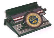 Photo of a stamped metal dark green Simplex 15 toy typewriter. It has a somewhat realistic angled grey keyboard with printed flat faux keys. Though the circular index is retained on this later model Simplex, a simple carriage moves the paper beneath the selected letter and advances after the letter has been pressed down onto the sheet.