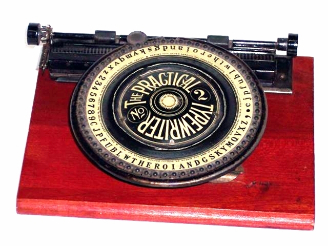 Simplex The Practical Typewriter Number 2. Depressing the dial moves the carriage one space. The dial is large, edged in black, with a beige center and large black lettering. There is a starburst in the middle of the dial. All upper and lower case letters are present, as well as numbers and figures. The wooden base is stained a thin red, allowing the grain to show through.