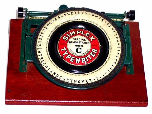 Simplex Special Demonstrated Model C Typewriter, upper and lower case with figs on a large dial. The body and paper feed assembly are painted green. There are two rubber paper feed rollers and two carriage twirlers. The large green dial has a wide, black inner ring around the logo plate. The logo plate has a red ring with the words SIMPLEX TYPEWRITER in sans serif caps. Inside the red ring, centered against a white circle, are the words SPECIAL DEMONSTRATED MODEL C. All the text is sans serif except the C, which is black and in caps. The much larger B is a large, bold, black serif B.