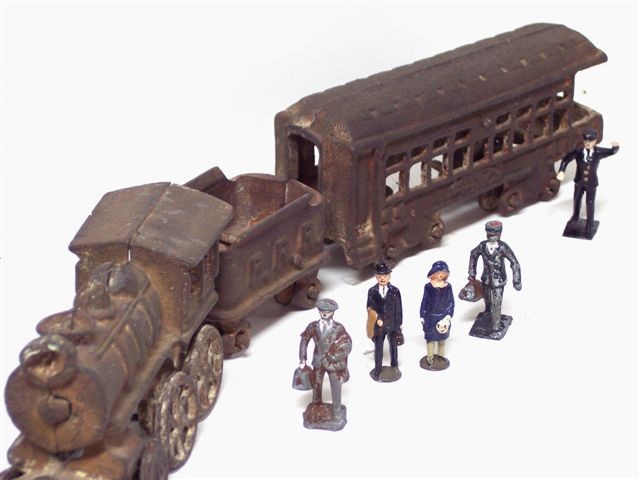 This picture includes a cast metal train with engine, tender, and passenger car having a platform at the rear. There are five figures. One is a young fellow in a brown suit, sporting a nice driver's cap, and carrying a brown portmanteau. Just behind him, a gentleman in a blue suit and white shirt with tie, wearing a hat, carrying a tan coat over one arm, and a doctor's bag in hand. To his left is a woman in a blue empire style dress over a pink blouse, wearing a blue stylish hat, white stockings, and carrying a small handbag. Behind her is a porter in black uniform over a white shirt, wearing a bow tie and a white porter's cap, and carrying a small suitcase. Back at the end of the train a uniformed conductor has one arm raised, perhaps signalling the train engineer.