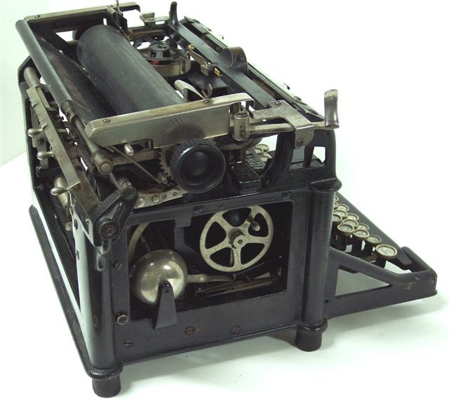 Here we have a view of the left side of the Underwood 1, taken from slightly above so as also to show the ribbon spools in place, and the 3-position line spacing selector at the rear of the carriage left of the paper tray. Below is the large bell and the other ribbon winding wheel.
