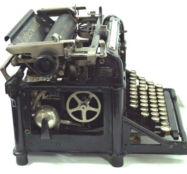 This photo of the left side of the typewriter shows its attractive yet simple design, with the keyboard frame and its supporting buttress, the large open window exposing the inner works, the columnar design of each corner of the typewriter, the thin skirting around the bottom of the frame, and the curved top front of the frame.